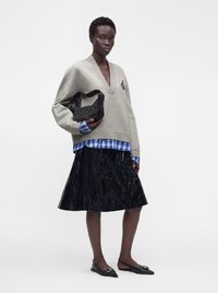 Femme portant un pull gris oversize par-dessus une chemise à carreaux bleue, une jupe noire texturée, des ballerines noires pointues, tenant un sac noir clouté.
