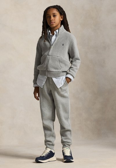 Junge Person mit langen Haaren, die einen grauen Sweatjacke mit Reißverschluss, graue Jogginghosen, ein gestreiftes Shirt und marineblaue und weiße Turnschuhe trägt, steht vor einem schlichten Hintergrund.