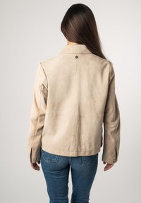 Beige Wildlederjacke mit kurzem Design, genähten Details auf der Rückseite und Druckknopfverschluss am Kragen. Kombiniert mit blauen Jeans.