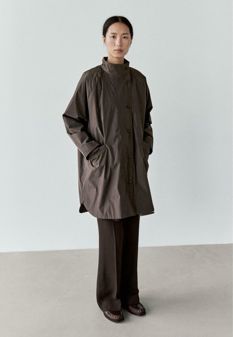 Brązowy pikowany płaszcz z wysokim kołnierzem, zapinany na guziki z bocznymi kieszeniami, zestawiony z ciemnymi spodniami i teksturowanymi butami. Gładkie, matowe wykończenie.