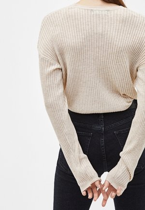 Person trägt einen beigen, gerippten Langarmpullover und hoch taillierte schwarze Jeans, mit verschränkten Händen auf dem Rücken.