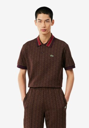 Joven con cabello negro corto que lleva una camisa polo marrón con estampado y pantalones a juego con logo verde de cocodrilo.