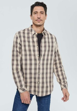 Chemise à carreaux beige et noire, à manches longues, en tissu mélangé coton, col standard, coupe droite, portée sur un t-shirt noir.