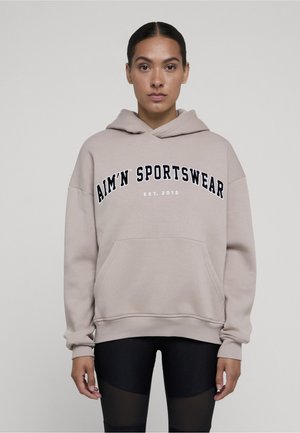 Femme aux cheveux foncés portant un sweat à capuche beige avec l'inscription "AIM'N SPORTSWEAR EST. 2013" se tenant devant un fond gris clair uni.