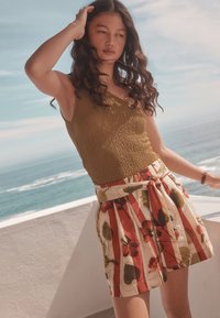 Débardeur en maille vert olive associé à une jupe plissée à motif floral avec des rayures rouges et crème. La lumière du soleil met en valeur la texture du tissu.