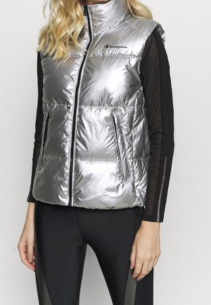 Kobieta ubrana w błyszczącą srebrną pikowaną kamizelkę na czarnym, długim, siateczkowym topie i czarnych legginsach, stojąca na jednolitym tle.