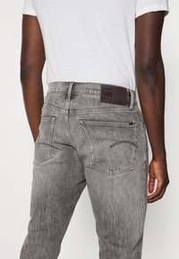 Jeans de denim gris con un acabado deslavado, que cuentan con un parche de cuero marrón etiquetado "RAW" y dos bolsillos traseros con detalles de costura curva.