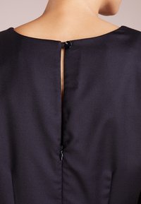 Černé šaty s hladkým materiálem, s detaily v oblasti zadního výstřihu, které jsou uzavřeny kulatým knoflíkem a skrytým zipem pro zapínání.