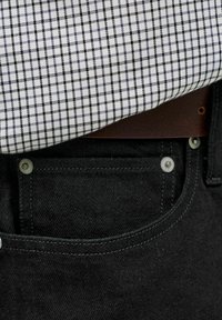 Schwarze Jeanshose mit silbernen Nieten, einem braunen Ledergürtel und einem in die Hose gesteckten weißen Hemd mit schwarzem Karomuster.