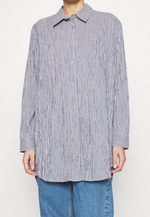 Langarm blouse met verticale grijs-witte strepen, gedragen over een blauwe spijkerbroek, vooraanzicht met handen ontspannen langs het lichaam.