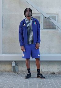 Sportinis aprangos derinys su mėlyna Adidas striuke, dryžuota palaidine, mėlynais šortais su Italijos logotipu ir juodais batais su baltais akcentais.