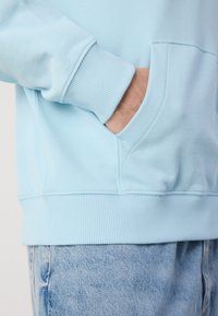 Sudadera azul claro con puños y dobladillo acanalados, que presenta un bolsillo delantero y una textura suave, combinada con jeans de denim azul claro.