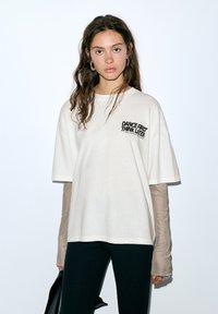 Ljusbeige oversize t-shirt med svart text "DANCE FIRST THINK LATER" på bröstet. Tyget verkar mjukt; matchas med svarta leggings och en lager-på-lager långärmad tröja under.