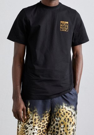 Persona che indossa una maglietta nera con un logo giallo sul petto e pantaloncini con motivo leopardato giallo e nero.