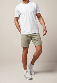 T-shirt bianca a maniche corte abbinata a pantaloncini kaki chiaro e sneakers alte bianche. L'outfit presenta un design semplice e colori neutri.