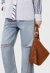Borsa in suede marrone con forma triangolare, parte superiore arrotondata e una larga tracolla con hardware dorato. Indossata con una camicia a righe e jeans strappati.