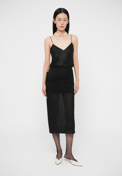 Czarna satynowa koszulka na cienkich ramiączkach, zestawiona z przezroczystą czarną ołówkową spódnicą z marszczeniami. Całość dopełniają białe buty na obcasie.