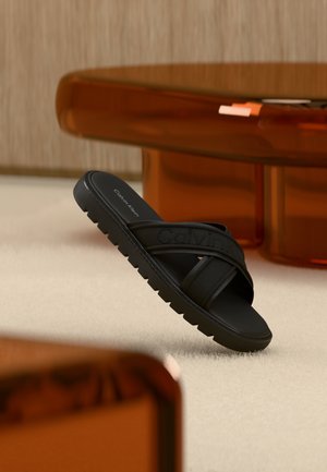 Sort Calvin Klein skyvesandal med teksturert såle som hviler på lyst teppe foran moderne møbler i ravfarge.