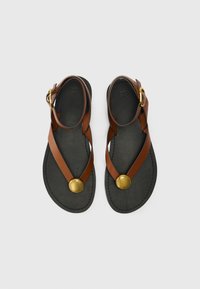 Sandali in pelle marrone con cinturini incrociati, accentato rotondo in oro e suola piatta nera. Fibbia regolabile alla caviglia per una vestibilità perfetta.