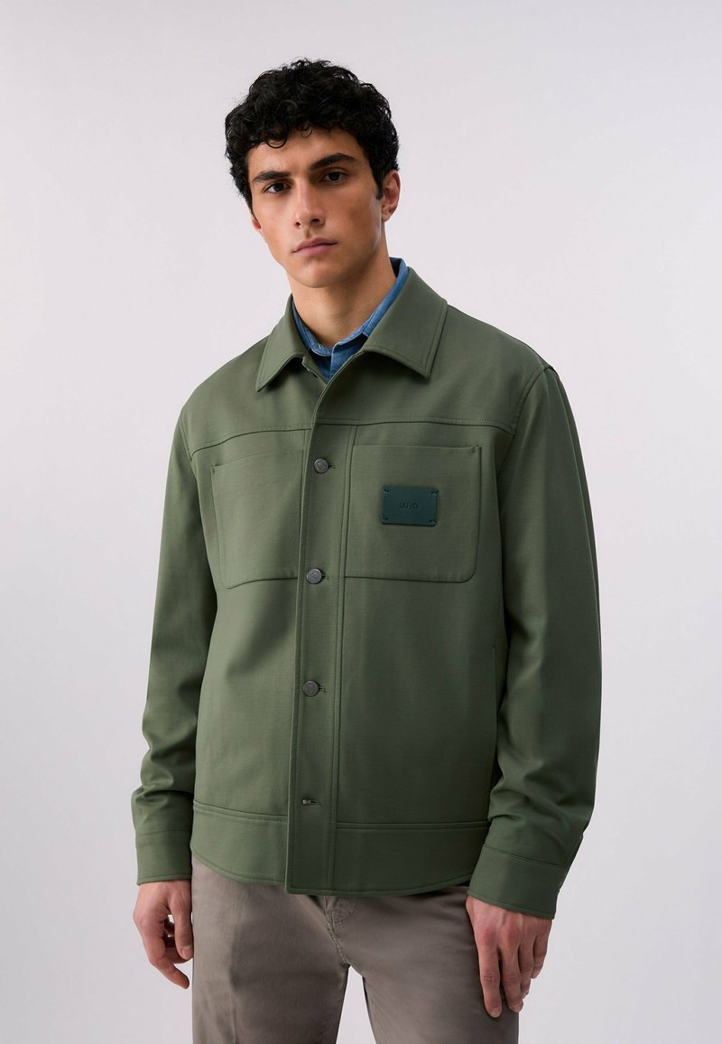Jeune homme portant une veste boutonnée vert olive avec poches poitrine sur une chemise bleue, debout devant un fond clair uni.