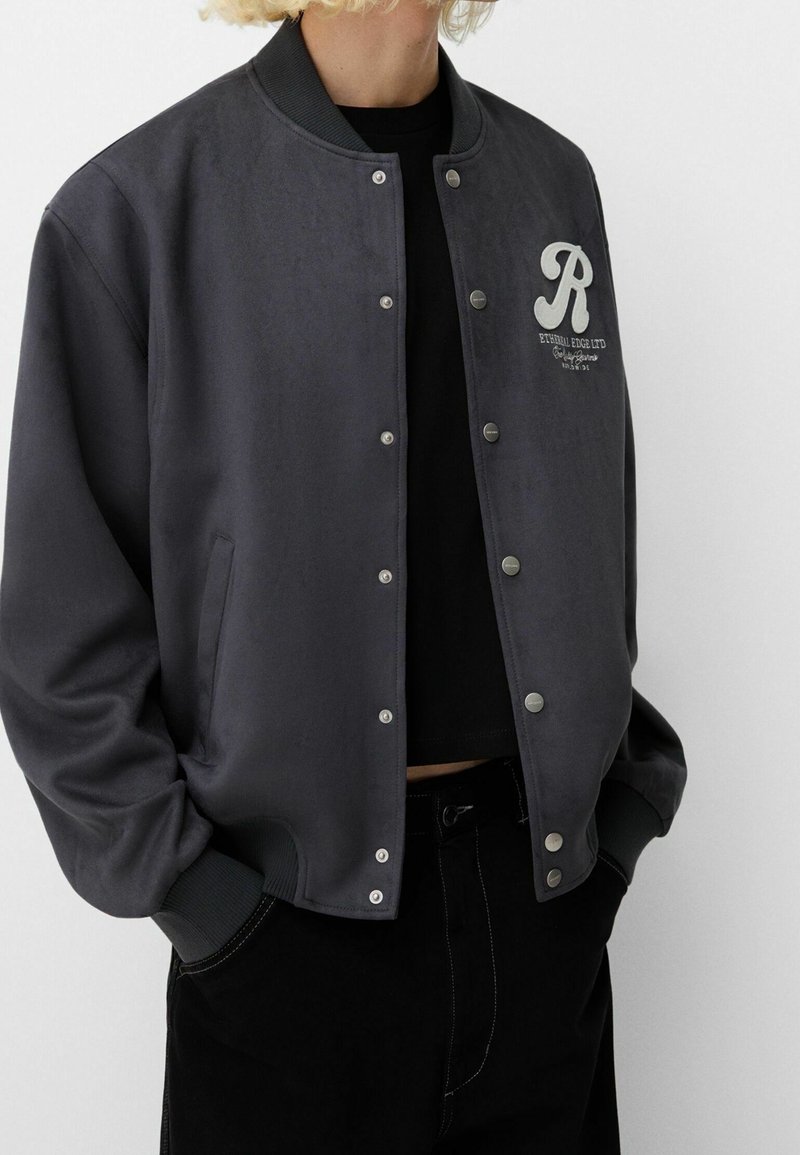 Gray varsity jacket with a smooth texture, ribbed collar, snap buttons, and a contrasting letter emblem on the chest. Features side pockets.