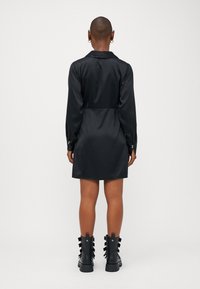 Vestido de cetim preto com mangas longas, corpete ajustado e saia ligeiramente godê. Apresenta punhos abotoados e um colarinho. Usado com botas pretas chunky.