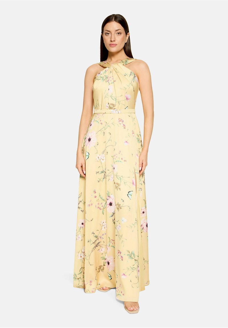 Femme portant une robe longue jaune à fleurs avec un col halter et un design sans manches, debout devant un fond uni.