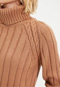 Pull à col roulé en maille torsadée de couleur fauve chaude, avec un côtelé texturé et un motif tressé distinctif sur l'épaule.