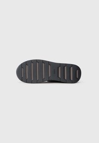 Semelle en caoutchouc noir de sneaker avec un motif texturé, présentant des sections surélevées alternées pour une meilleure traction et stabilité. Logo de la marque visible.