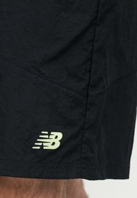 Μαύρα αθλητικά σορτς με ραμμένα ραφές και λευκό λογότυπο New Balance κοντά στο τελείωμα πάνω από το κατώτερο μέρος του ποδιού ενός ατόμου.