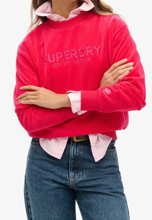 Osoba nosząca jaskraworóżową bluzę Superdry na jasnoróżowej koszuli z kołnierzykiem, w połączeniu z niebieskimi dżinsami, z rękami skrzyżowanymi.