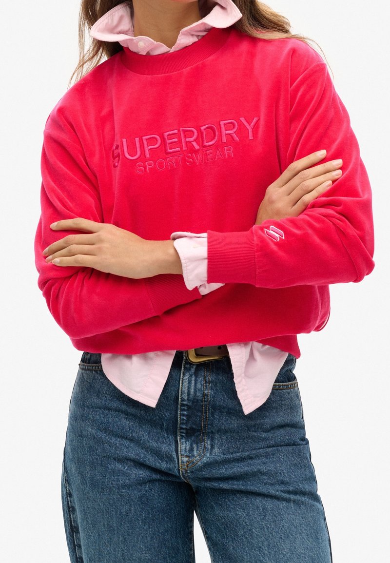 Personne portant un sweat-shirt Superdry rose vif sur une chemise à col rose pâle, associé à un jean bleu, bras croisés.