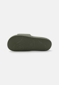 Olíva zöld lábbeli, texturált gumitalppal, amelyen kis kiemelkedő pöttyök vannak a tapadás érdekében, míg a felső része sima felületű. "Lacoste" felirattal ellátva.