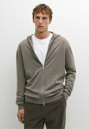 Giovane uomo che indossa una felpa grigia con zip sopra una camicia bianca e pantaloni scuri, in piedi con una mano in tasca davanti a uno sfondo bianco semplice.