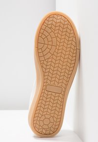 Semelle d'une chaussure en caoutchouc marron clair avec des motifs de bande de roulement circulaires et en chevrons, positionnée verticalement contre un coin blanc.