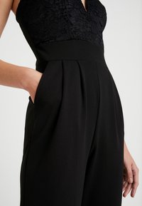 Kvinna iförd en svart jumpsuit med spetsöverdel, handen i fickan, som visar upp plisserade byxor och figursydd midjedetalj.