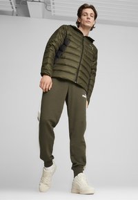 Olivengrøn pufferjakke med sorte sidepaneler, lynlås og hætte; parret med matchende joggingbukser med hvidt logo og sidekanter.