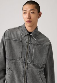 Γκρι τζιν μπουφάν με φερμουάρ στο μπροστινό μέρος, μυτερό γιακά και δύο τσέπες με καπάκι. Διαθέτει σχεδιασμό με ραμμένα πάνελ και μαλακό, υφασμάτινο ύφος με υφή.