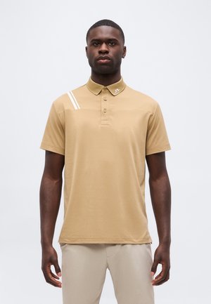 Homme portant un polo beige à manches courtes avec deux bandes blanches sur l'épaule et un pantalon clair, debout devant un fond uni.