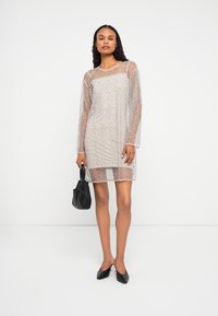 Nő áttetsző, hosszú ujjas, térd középig érő ruhát visel, amely alatt egy testhez simuló, nude színű alsóruha van, fekete kézitáskát tart a kezében, és fekete hegyes orrú cipőt visel.