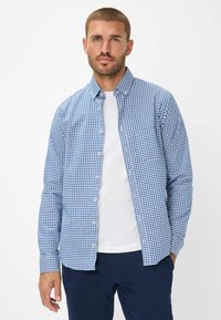 Camicia a quadri blu e bianchi, con maniche lunghe, colletto, realizzata in tessuto, indossata sopra una t-shirt bianca, abbinata a pantaloni scuri.