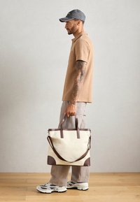 Torba typu tote z brązowymi akcentami, mocnymi uchwytami i odpinanym paskiem na ramię. Nosi się ją z teksturowaną beżową bluzką i jasnobeżowymi spodniami.