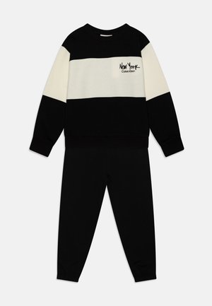 Completo di felpa e pantaloni in blocco di colore nero e crema. La felpa presenta un collo rotondo e un logo, abbinata a pantaloni neri a gamba affusolata con polsini elasticizzati. Composizione in miscela di cotone.