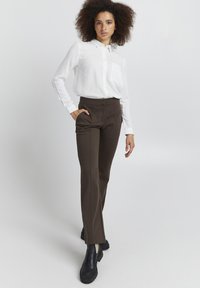 Biela košeľa s dlhým rukávom a vreckom, spárovaná s tmavohnedými nohavicami s vysokým pásom. Čierne robustné topánky dokončujú outfit.