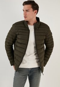 Olivengrøn puffjakke med zip foran, ribbet krave og vandret quiltet design. Båret over en hvid t-shirt med rund hals og parret med jeans.