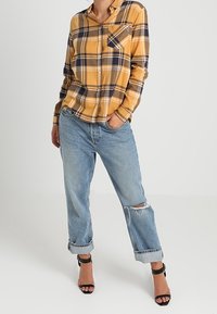 Camicia a quadri gialla e blu marino con bottoni, abbinata a jeans chiari strappati e tacchi neri con cinturini, che mettono in mostra un taglio rilassato e una tasca sul petto.