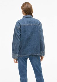 Denim jakna u tamnoplavoj boji, oversized dizajn s dugim rukavima i manžetama ukrašenim teksturiranim perlicama. Pogled s leđa prikazuje jednostavnu konstrukciju.