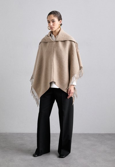Poncho en laine beige avec un ourlet frangé, dotée d'un large col et d'un design à devant ouvert. Porté sur un haut blanc et un pantalon noir.