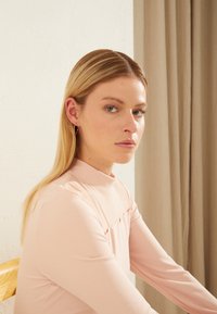 Top de manga longa em rosa pálido com um colarinho alto e detalhe de pregas subtil. Textura suave, design ajustado. Modelo sentado contra um fundo neutro.