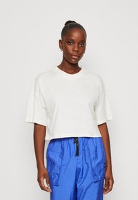 T-shirt bianco cropped con maniche corte e scollatura a V; presenta un logo beige discreto sul lato sinistro del petto; indossato con pantaloni blu lucidi.